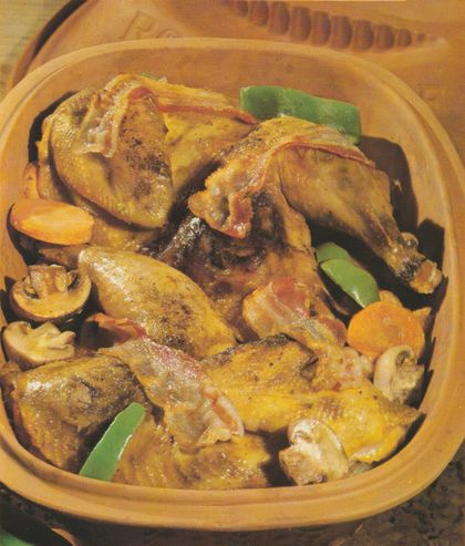 Gebratener Fasan in goldbrauner, glänzender Haut, gevierteilt in einem runden Römertopf, garniert mit Champignonscheiben und Stücken grüner Paprika. Das Geflügel liegt auf einem Bett aus geschmortem Gemüse mit Zwiebeln und Möhrenscheiben.