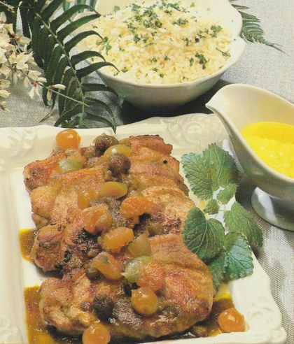 Gebratene Kalbsrückensteaks mit glasierten Früchten aus dem „Rüdesheimer Früchtetopf”. Daneben eine Saucière mit gelber Holländischer Soße und im Hintergrund eine Schüssel mit Reis, bestreut mit frischen Kräutern. Dekoriert mit einem Farnzweig und weißen Blüten.