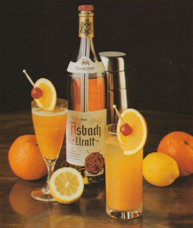 Eine Flasche Asbach Uralt steht neben einem Cocktail-Shaker und zwei gefüllten Gläsern mit orangefarbenen Drinks, die jeweils mit einer Orangenscheibe und Cocktailkirsche garniert sind. Im Hintergrund liegen frische Orangen und Zitronen auf dunklem Untergrund.