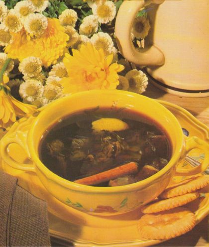 Eine dunkle Fleischbrühe mit Karotten, Pilzen und einem Klecks Butter in einer gelben Suppenterrine mit Blumenmuster. Im Hintergrund stehen gelbe und weiße Blumen in einer Vase sowie eine passende Tasse auf einem Unterteller.