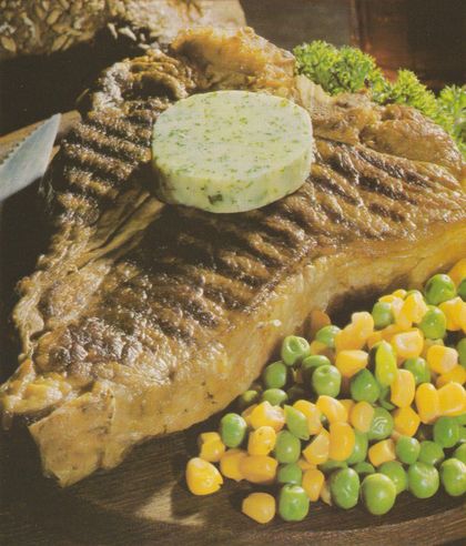 Gegrilltes T-Bone-Steak mit deutlichen Grillstreifen auf einem Teller, darauf eine runde Scheibe Kräuterbutter. Als Beilage Erbsen-Mais-Gemüse und im Hintergrund krause Petersiliengarnitur.