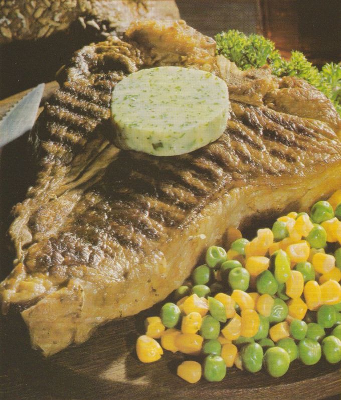 Gegrilltes T-Bone-Steak mit deutlichen Grillstreifen auf einem Teller, darauf eine runde Scheibe Kräuterbutter. Als Beilage Erbsen-Mais-Gemüse und im Hintergrund krause Petersiliengarnitur.