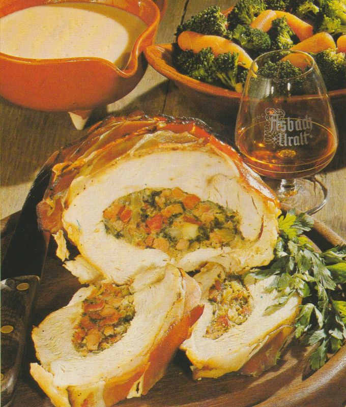 Gefüllte Putenbrust mit Schinkenspeck umwickelt, in Scheiben aufgeschnitten auf einem Holzbrett. Die Füllung besteht aus gewürfeltem Schinken, Zwiebeln und Petersilie. Im Hintergrund ein Schwenker mit der Aufschrift „Asbach Uralt”, eine Schüssel mit Brokkoli und Karotten sowie eine Terrine mit Sauce. Petersilie als Garnitur.