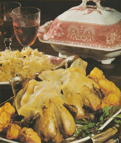 Goldbraun gebratene Fasane auf einer Platte, garniert mit frischer Petersilie und gelben Blüten. Im Hintergrund eine weißen Terrine mit rotem Dekor sowie ein Glas Weißwein. Links daneben Mandelkroketten auf einem separaten Teller.