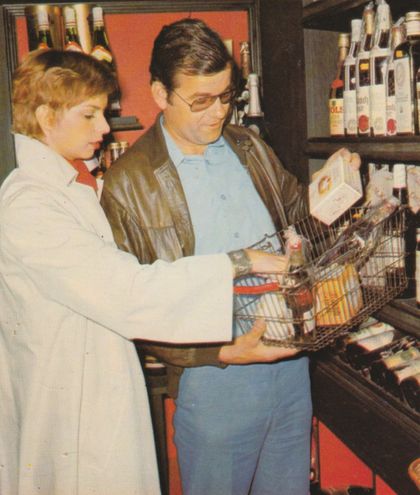 Ein Kunde steht im Supermarkt vor einem Regal mit Spirituosen und betrachtet gemeinsam mit der Verkäuferin einen gefüllten Einkaufswagen mit verschiedenen Lebensmitteln und Verpackungen. Im Hintergrund sind Flaschen auf einem Regal sowie Dekoration an der Wand zu erkennen.