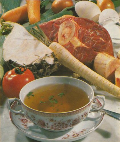 Eine Tasse klare Fleischbrühe mit Petersilie steht im Vordergrund, umgeben von frischem Suppengemüse wie Sellerie, Möhren, Pastinaken, Porree, einer Tomate und einem rohen Fleischstück. Im Hintergrund sind weitere Zutaten zu sehen.