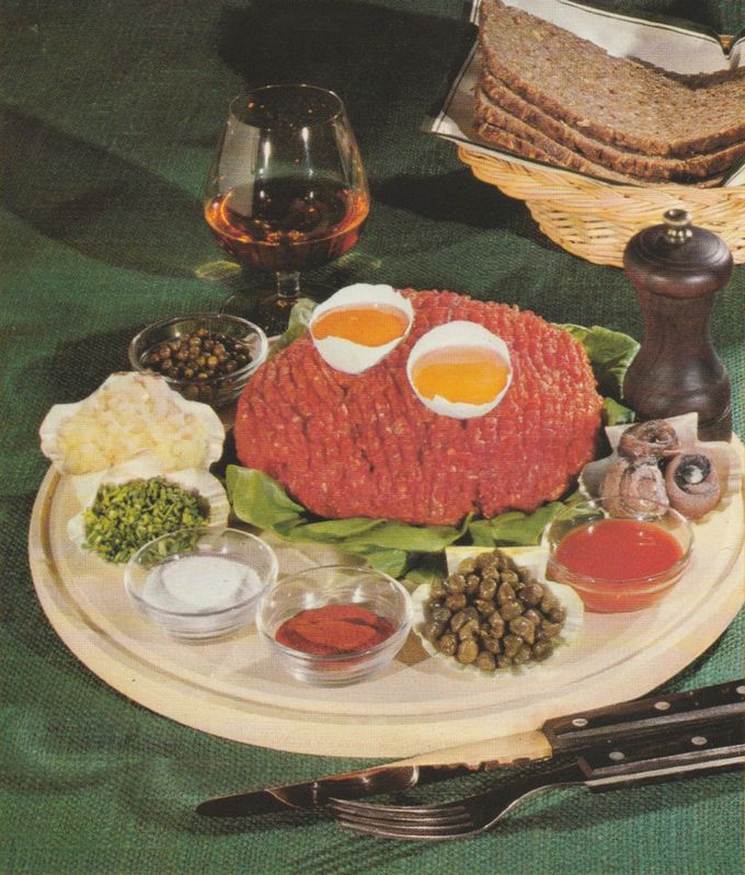 Rohes Beefsteak-Tatar auf einem Salatblatt, garniert mit zwei halbierten Eiern. Drumherum sind die Zutaten in kleinen Schälchen arrangiert: gehackter Schnittlauch, Salz, Paprikapulver, Tomaten-Ketchup, Kapern, gehackte Zwiebeln, schwarzer und grüner Pfeffer, Sardellen. Im Hintergrund ein Cognacschwenker mit Asbach Uralt, ein Brotkorb mit dunklem Brot und eine Pfeffermühle.