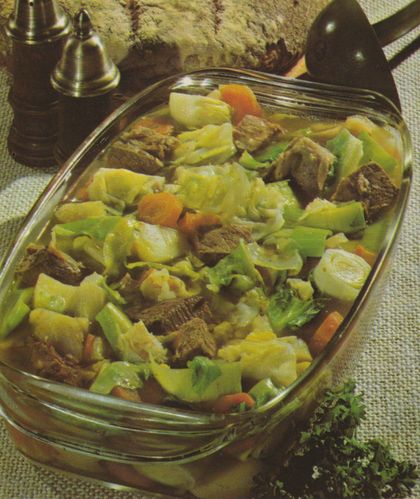 Ein herzhafter Eintopf mit großen Fleischstücken, Weißkohl, Karotten, Lauch und Kartoffeln in klarer Brühe in einer rechteckigen Glasform. Im Hintergrund stehen Salz- und Pfefferstreuer sowie ein Brotlaib auf karierter Tischdecke.