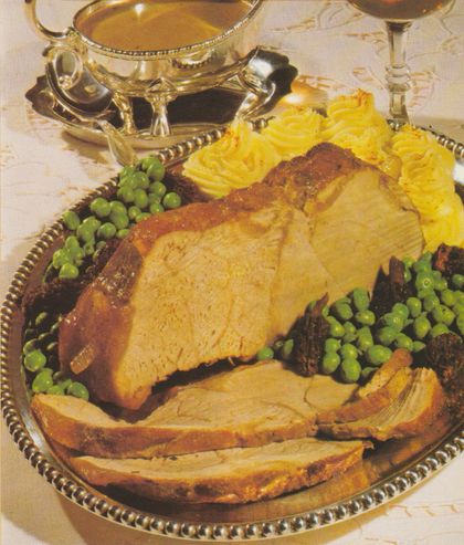 Gebratener Kalbsbraten auf einer silbernen Servierplatte, in Scheiben geschnitten, umgeben von grünen Erbsen und Möhrenstücken, garniert mit gespritztem Kartoffelpüree. Im Hintergrund eine goldene Sauciere auf weißer Spitzendecke.