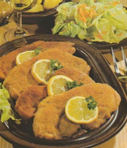 Vier panierte Kalbsschnitzel auf braunem Teller, jeweils mit einer Zitronenscheibe und Petersilie garniert. Auf einem Holzbrett serviert mit Salatbeilage aus Kopfsalat und geraspelten Möhren. Im Hintergrund Weinglas und Besteck.