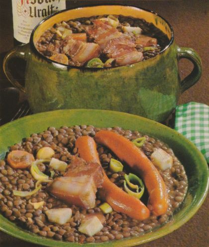 Ein herzhafter Linseneintopf mit Würstchen, Möhrenscheiben, Porree und Speckstücken wird in einem grünen Teller im Vordergrund und einem grün-gelben Schmortopf im Hintergrund serviert.