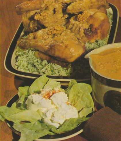 Gebratene Hähnchenstücke auf Petersilienreis in einer dunklen Platte, daneben eine Schüssel mit Nusssauce. Im Vordergrund ein Teller mit Frischkäse auf Salatblättern, mit Paprikapulver bestreut.