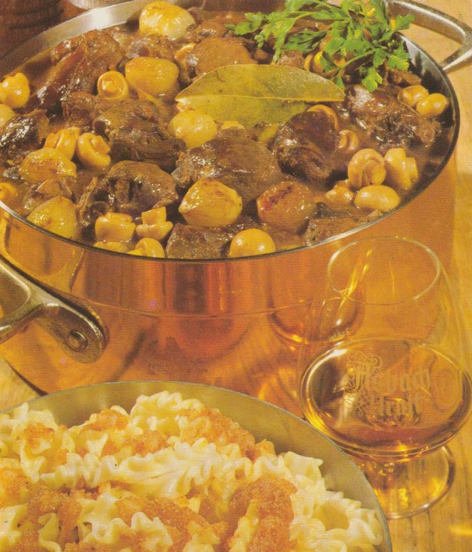 Kupferfarbener Schmortopf mit Hasenpfeffer: dunkle Fleischstücke in brauner Sauce mit Champignons, kleinen Zwiebeln, Nudeln und Lorbeerblatt. Daneben ein Glas Bier und ein Teller mit Butternudeln, bestreut mit geröstetem Zwiebackmehl.