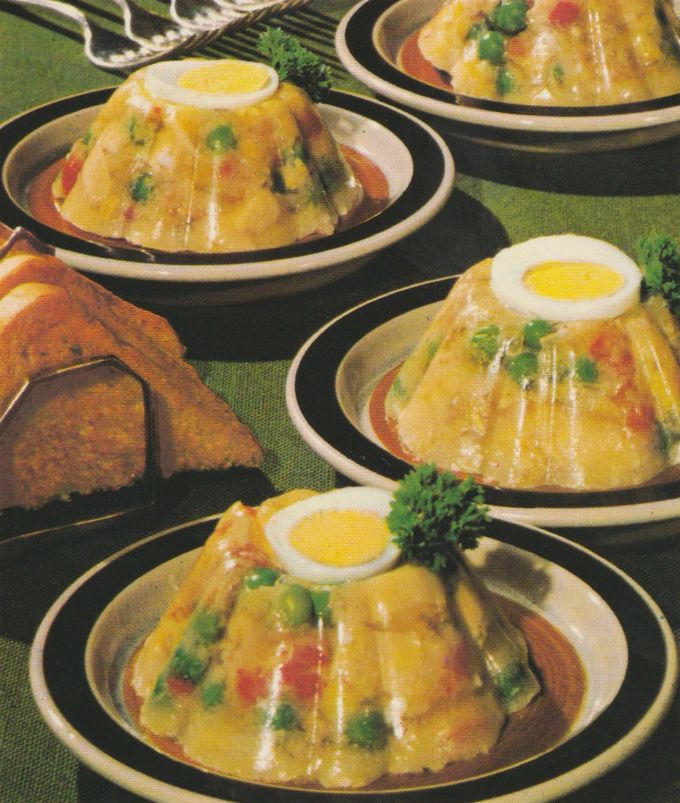 Kuppelförmige Gelee–Sulzen aus Hähnchenfleisch mit Erbsen und Paprikawürfeln, überzogen mit glänzender Aspikschicht. Jede Portion ist mit einer halben Eischeibe und Petersilie garniert und wird auf Tellern mit dunklem Rand serviert. Im Hintergrund Toastbrot auf einem Holzbrett.