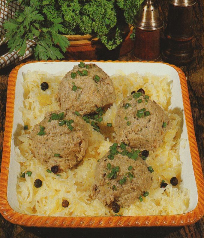 Vier panierte Fleischbällchen liegen auf einem Bett aus Sauerkraut, garniert mit gehacktem Schnittlauch und schwarzen Pfefferkörnern. Im Hintergrund befinden sich frische Petersilie in einem Topf, ein kariertes Geschirrtuch sowie Salz- und Pfefferstreuer aus Holz.