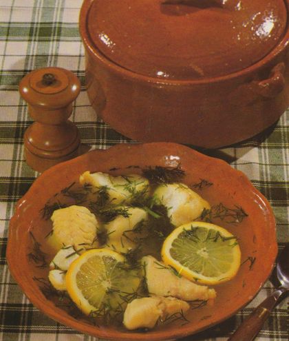 Ein brauner Tonteller mit gekochten Kartoffeln und Fischstücken, garniert mit Zitronenscheiben und frischem Dill in Brühe. Im Hintergrund stehen ein brauner Tontiegel mit Deckel, eine Pfeffermühle aus Holz und ein kariertes Tischtuch in Grün und Beige.