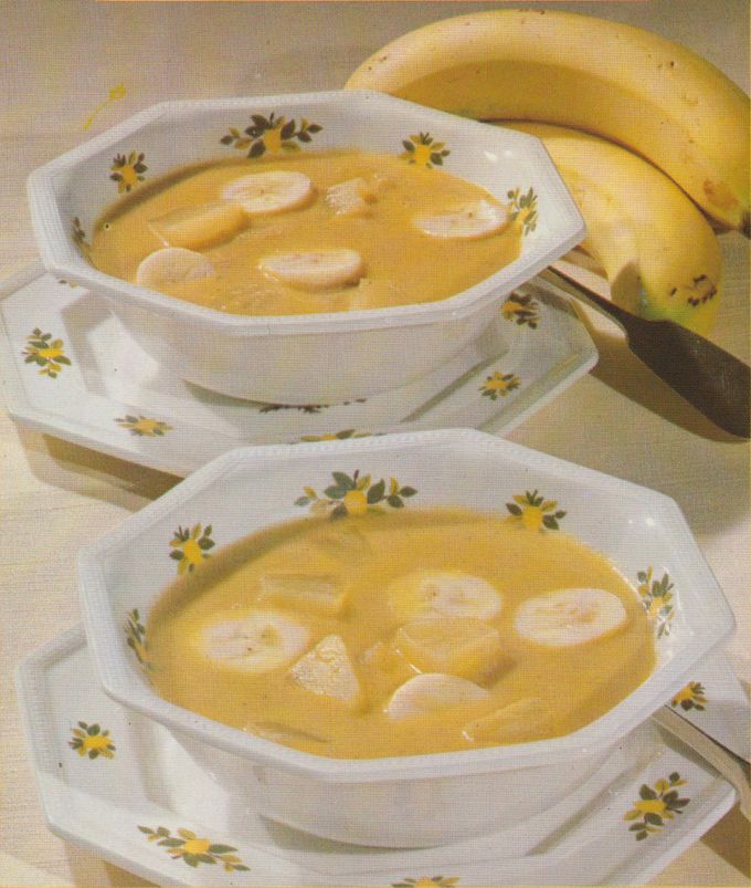 Eine cremige gelbe Bananensuppe mit Bananenscheiben in zwei weißen Schüsseln mit gelbem Blumenmuster, daneben liegen frische Bananen. Die Suppenschüsseln stehen auf passenden Untertellern mit demselben Dekor.