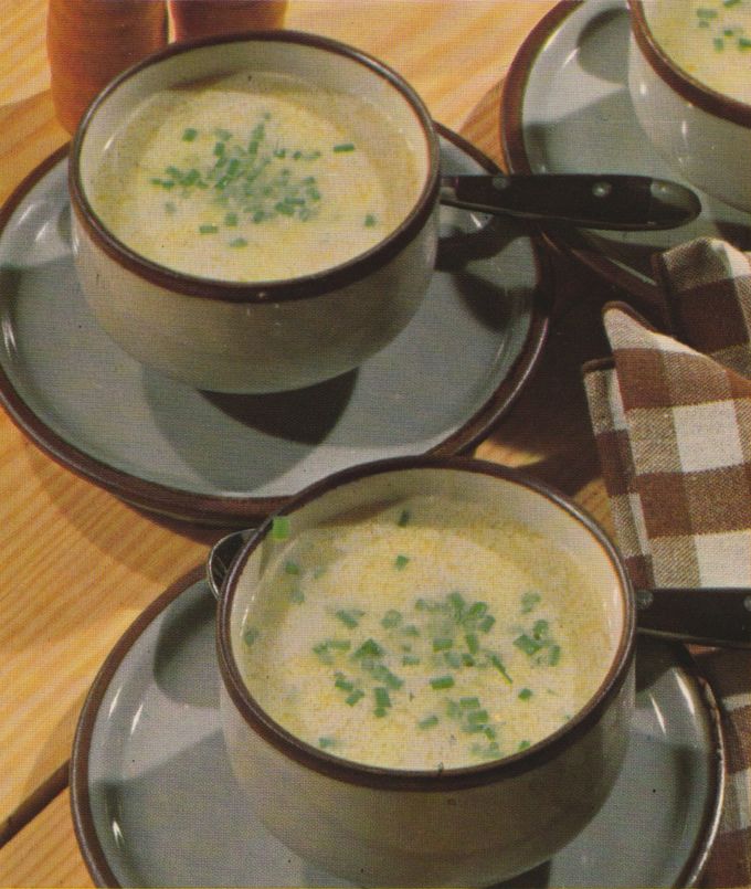 Zwei Suppentassen mit cremiger, heller Suppe, garniert mit gehackten Kräutern, stehen auf Untertassen neben einer karierten Serviette und sind auf einem hellen Holztisch arrangiert.