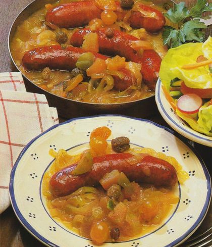 Geräucherte Mettwürstchen in einer Pfanne mit gedünsteten Zwiebelringen, Aprikosen, Pflaumen und Kapern in glänzender Soße. Im Vordergrund ein angerichteter Teller mit einer Portion des Gerichts auf einem Bett aus Sauerkraut. Rechts daneben ein kleiner Salat mit Radieschen, Gurke und Petersilie. Kariertes Geschirrtuch als Unterlage.