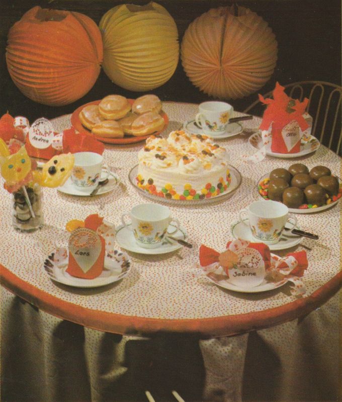 Ein festlich gedeckter Tisch mit einer Sahnetorte in der Mitte, umgeben von Tassen mit Blumenmuster, Gebäck und Schokoladenkugeln. Papier–Lampions hängen darüber, und Platzdekorationen mit Papp-Bonbons und Namensschildern schmücken die einzelnen Gedecke.