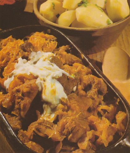 Schweinegulasch mit Sauerkraut in einer dunklen Pfanne, mit einem Klecks saurer Sahne belegt. Im Hintergrund eine Schüssel mit Salzkartoffeln sowie ein Holzlöffel.