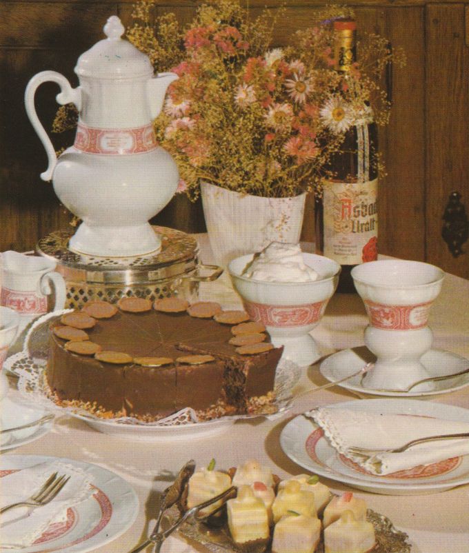 Eine festliche Kaffeetafel mit Schokoladentorte, die mit Marzipanplättchen dekoriert ist, weißem Porzellan mit rosafarbenem Dekor, einer Flasche Asbach Uralt und einem Strauß Trockenblumen. Im Vordergrund steht ein Teller mit Spargel in heller Sauce.