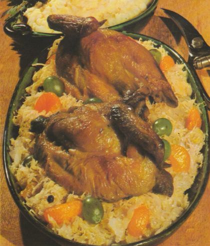 Gebratene Hähnchenhälften auf einem Bett aus Weinkraut, garniert mit Mandarinenstücken, halbierten Weintrauben und Wacholderbeeren, serviert in einer ovalen Auflaufform. Im Hintergrund Kartoffelpüree in einer Schüssel.