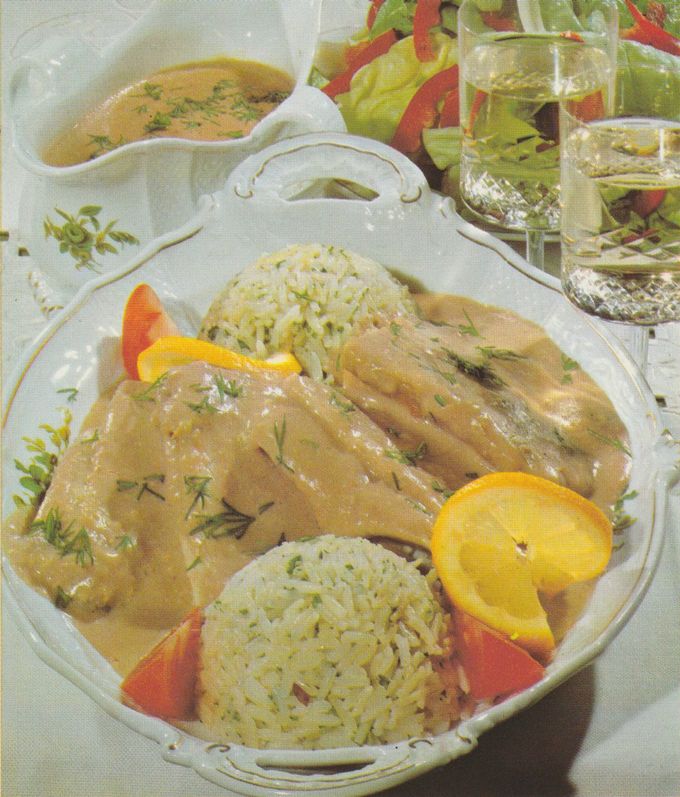 Schollenfilets in Sauce auf einem ovalen Teller mit dekorativem Goldrand, garniert mit Dill und Zitronenscheiben. Dazu zwei Portionen Petersilienreis und Tomatenstücke. Im Hintergrund eine Sauciere mit Sauce, ein Salat aus Kopfsalat und Paprika sowie Wassergläser.