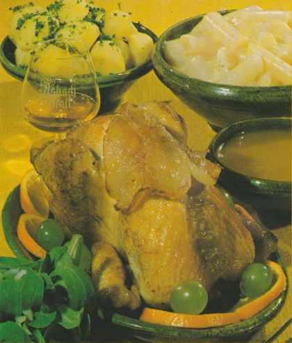 Gebratenes Perlhuhn auf einer Platte, garniert mit Orangenscheiben, grünen Weinbeeren und frischer Petersilie. Im Hintergrund Schüsseln mit Kartoffeln und Schwarzwurzelgemüse sowie ein Cognacschwenker mit Asbach Uralt und eine Sauciere.