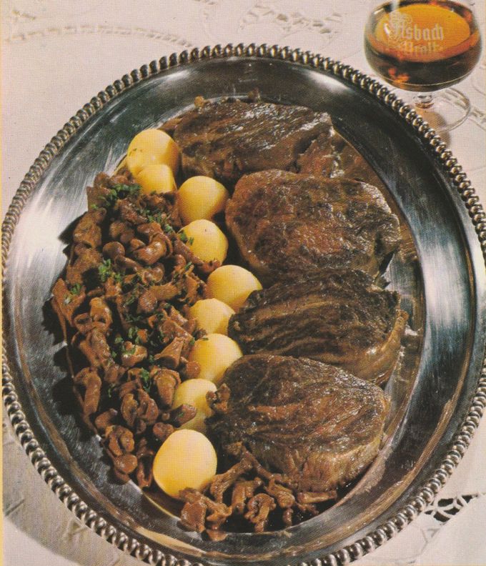 Gebratene Filetsteaks auf einer silbernen Servierplatte mit Perlrand, dazu gedünstete Pfifferlinge mit Petersilie und runde Kartoffelbällchen. Im Hintergrund ein Glas Asbach Uralt.