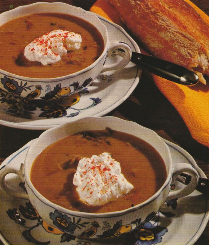 Zwei Suppentassen mit cremiger, orangebrauner Suppe, garniert mit Sahnehaube und Paprikapulver, stehen auf Untertassen mit Vogelmotiv. Daneben liegt knuspriges Weißbrot mit Messer auf einem Holzbrett.