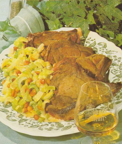 Vier gebratene Frischlingskoteletts auf einem weißen Teller mit grünem Blumenmuster, dazu Staudensellerie-Gemüse mit Erdnüssen. Im Vordergrund ein Glas Asbach Uralt, im Hintergrund grün-weißes Geschirrtuch und frische Salatblätter.