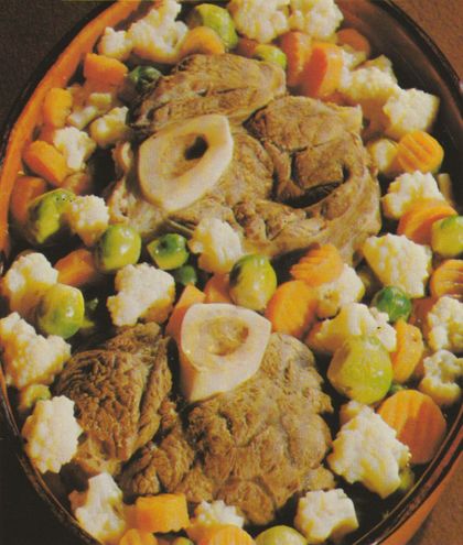 Gebratene Ochsenbeinscheiben mit Knochenmark in einer ovalen Schüssel, umgeben von gedünstetem Gemüse: Möhren, Blumenkohl, Rosenkohl und Erbsen. Das Knochenmark ist in der Mitte der Fleischscheiben sichtbar.