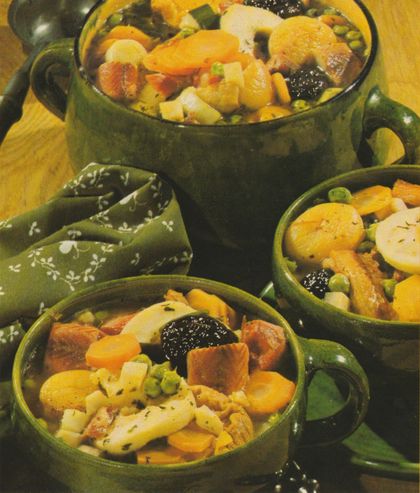 Drei olivgrüne Keramikschalen gefüllt mit einem deftigen Eintopf aus Kartoffeln, Möhren, Erbsen, Fleischstücken und Trockenfrüchten, arrangiert auf gelber Stoffserviette mit Blumenmuster.