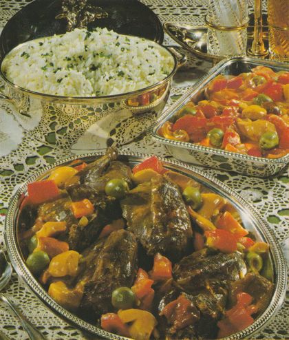 Drei silberfarbene Servierplatten auf gehäkelter Tischdecke: vorne gebratene Rebhühner mit Paprikawürfeln, Steinpilzen und grünen Oliven in Sauce, dahinter rechts ein ähnliches Gericht in heller Sauce, links eine Messingschale mit Reis, bestreut mit gehackten Kräutern. Im Hintergrund Glaskaraffen.
