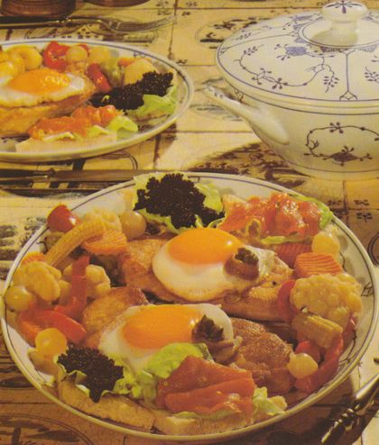 Zwei reich garnierte Kalbsschnitzel auf Tellern mit blauem Blumenmuster, serviert auf rustikaler Holzunterlage. Jedes Schnitzel ist mit einem Spiegelei, Sardellenfilets und Kapern belegt. Daneben jeweils geröstetes Toastbrot mit Salatblatt, Kaviar und Räucherlachs. Als Beilagen Röstkartoffeln, Mixed Pickles, Blumenkohl und kleine Maiskolben. Im Hintergrund eine weiße Suppenterrine mit blauem Dekor.