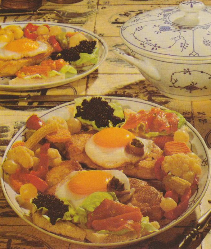 Zwei reich garnierte Kalbsschnitzel auf Tellern mit blauem Blumenmuster, serviert auf rustikaler Holzunterlage. Jedes Schnitzel ist mit einem Spiegelei, Sardellenfilets und Kapern belegt. Daneben jeweils geröstetes Toastbrot mit Salatblatt, Kaviar und Räucherlachs. Als Beilagen Röstkartoffeln, Mixed Pickles, Blumenkohl und kleine Maiskolben. Im Hintergrund eine weiße Suppenterrine mit blauem Dekor.