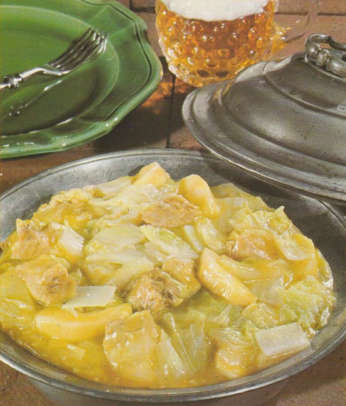 Eintopf aus geschmortem Schweinebauch, Chinakohl und Apfelscheiben in gelblich-goldener Curry-Soße, serviert in einer ovalen Zinn-Schüssel mit abgehobenem Deckel. Im Hintergrund grüne Teller mit Gabel sowie ein bernsteinfarbenes Hobnail-Glas auf einem Holztisch.