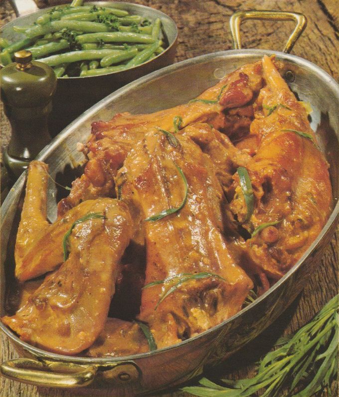 Glasierte Kaninchenstücke in einer ovalen Bratform, garniert mit frischen Estragonzweigen. Die goldbraun gebratenen Fleischteile glänzen von der Sahne-Sauce. Im Hintergrund eine Schüssel mit grünen Bohnen und ein Pfefferstreuer auf geflochtener Unterlage.