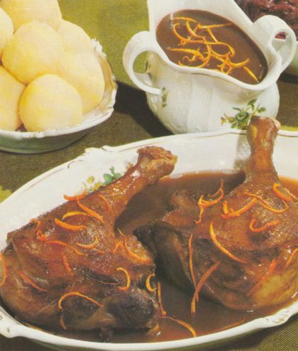Gebratene Ente auf ovalem Teller, garniert mit feinen Orangenzesten-Streifen. Daneben eine Sauciere mit dunkler Sauce, ebenfalls mit Orangenzesten verziert. Im Hintergrund eine Schale mit Salzkartoffeln.