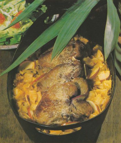 Schwarze Schmorpfanne mit überbackenen Schweinesteaks auf einem Bett aus Bandnudeln, Apfelschnitzeln und Zwiebeln. Das Fleisch ist mit geriebenem Käse bestreut. Lange grüne Blätter einer Topfpflanze ragen dekorativ über die Pfanne. Im Hintergrund links ein Teller mit gemischtem Salat.