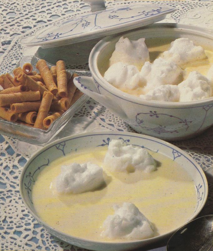 Vanillepudding mit Schneeklößchen auf einem Teller und in einer Schüssel, daneben goldbraune Waffelröllchen auf einem Glasteller. Das Geschirr mit blauem Blumenmuster steht auf einer weißen Häkeldecke.