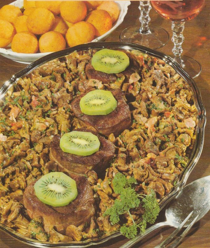 Ovale Servierplatte mit gebratenen Rehmedaillons, die jeweils mit einer Kiwi-Scheibe belegt sind, umgeben von Pfifferlingen in Rahmsauce mit Petersilie. Im Hintergrund eine weiße Schale mit goldgelben Kartoffelbällchen und Kristallgläser.