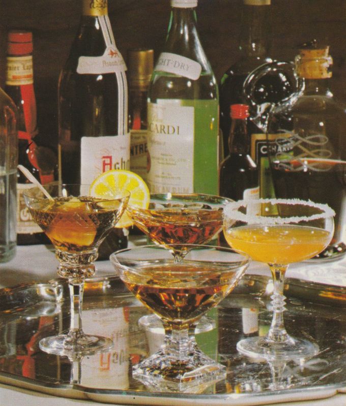 Eine Auswahl verschiedener Cocktails in Cocktailgläsern, darunter Drinks mit Zitronenscheibe und Zuckerrand, arrangiert vor einer Sammlung von Spirituosenflaschen wie Wermut, Wein und Likören. Die Gläser stehen auf einem spiegelndem Tablett, im Hintergrund sind verschiedene Flaschen Spirituosen.