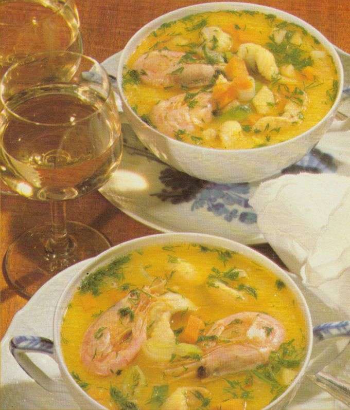 Zwei Suppentassen mit gelber Fischsuppe, die Lachsstücke, weiße Bohnen und frischen Dill enthält, serviert auf Untertellern mit blauem Blumenmuster. Daneben steht ein Weinglas mit Weißwein und eine weiße Stoffserviette auf einem Holztisch.