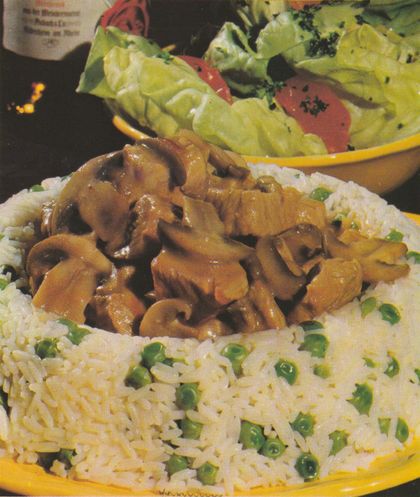 Geschmorte Champignons mit Kalbfleisch in cremiger Sauce in einem Reisrand mit Erbsen, serviert auf einem weißen Teller. Im Hintergrund eine gelbe Schale mit Kopfsalat.