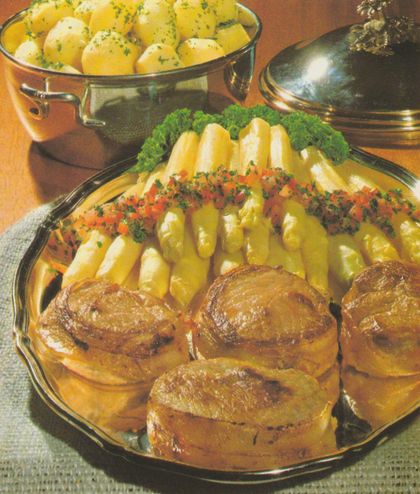 Vier gebratene Kalbsmedaillons mit Speckmantel auf einer ovalen Servierplatte, dahinter weißer Stangenspargel mit gehackten Tomaten und Petersilie, umrahmt von frischen Petersilienbüscheln. Im Hintergrund ein Topf mit Petersilienkartoffeln und ein silberner Deckel.