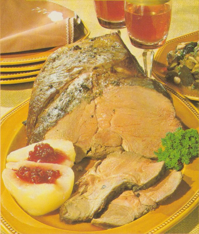 Gebratene Hirschkeule auf gelbem Serviertablett, in Scheiben aufgeschnitten, mit knuspriger Kruste und Kräutern. Daneben halbierte Birnen mit Preiselbeerfüllung und frische Petersilie als Garnitur. Im Hintergrund gestapelte gelbe Teller, ein Glas Getränk und eine Sauciere auf kariertem Geschirrtuch.