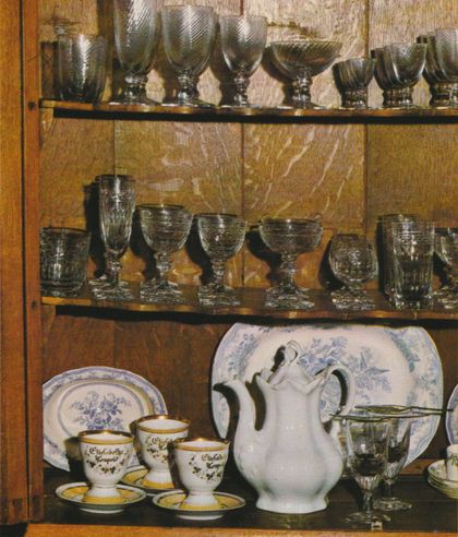 Ein Küchenschrank aus Holz mit drei Regalebenen zeigt Gläser und Geschirr: oben rauchfarbene Kelchgläser, in der Mitte verschiedene gemusterte Trinkgläser, unten weiße Porzellantassen mit goldener Beschriftung, ein Tablett mit blauem Blumenmuster und eine weiße Kaffeekanne.