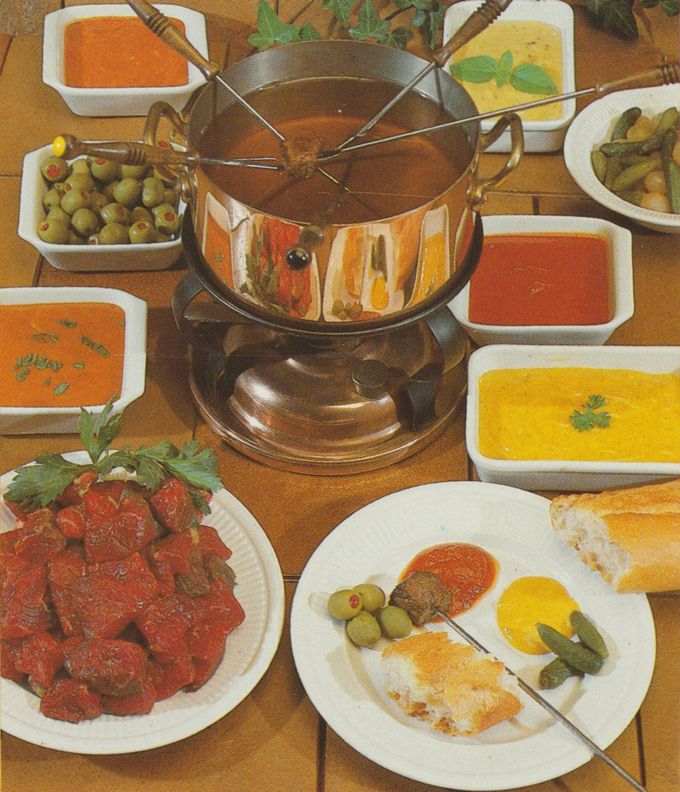 Fondue-Set aus Kupfer mit Fonduegabeln auf Holztisch, umgeben von weißen Schälchen mit verschiedenen Saucen in Orange und Gelb. Dazu grüne Oliven, eingelegte Gurken, Weißbrotstücke und ein angerichteter Teller mit Fleisch, Oliven, Gurken und Saucen.
